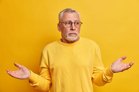 Portrait of puzzled hesitant bearded mature man spreads palms and faces difficult choice looks uncertain wears casual jumper optical glasses has confused expression isolated over yellow background Portrait of puzzled hesitant bearded mature man spreads palms and faces difficult choice looks uncertain wears casual jumper optical glasses has confused expression isolated over yellow background